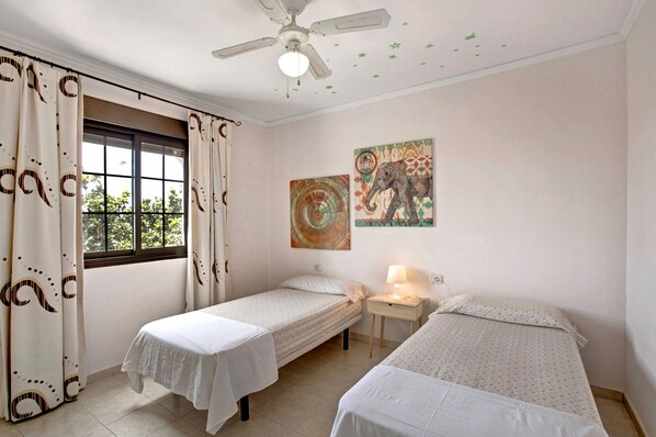 A room with two single beds, a ceiling fan, a window with curtains, and a wall with a decorative elephant painting.