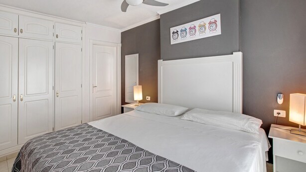 Habitación de hotel con cama, ventilador de techo, mesita de noche con lámpara, reloj de pared y armario.
