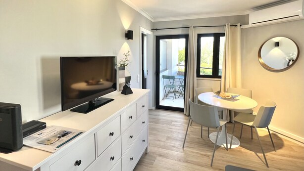 Ein modernes Wohnzimmer mit Flachbildfernseher, einer weißen Kommode, einem Esstisch mit Stühlen und einem Balkon mit Blick auf Grünflächen.