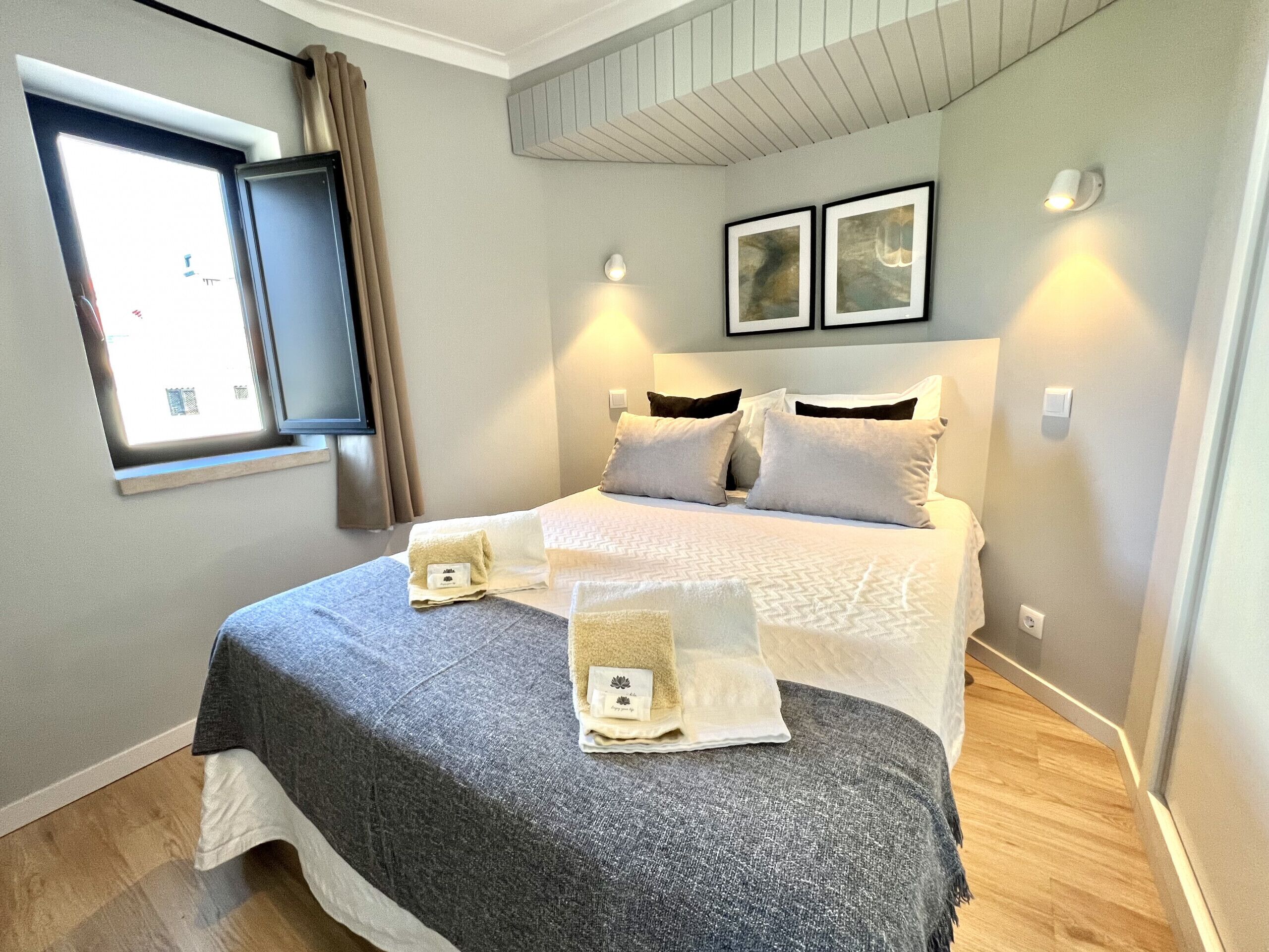 A neatly arranged bedroom with a bed, pillows, a bedside table, a window with curtains, and wall-mounted lights.