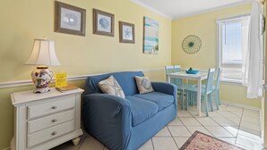 Huoneessa on sininen sohva, valkoinen yöpöytä, jossa on lamppu, sekä ruokapöytä tuoleineen. Seinällä on kehystettyjä kuvia ja ikkunassa sälekaihtimet.