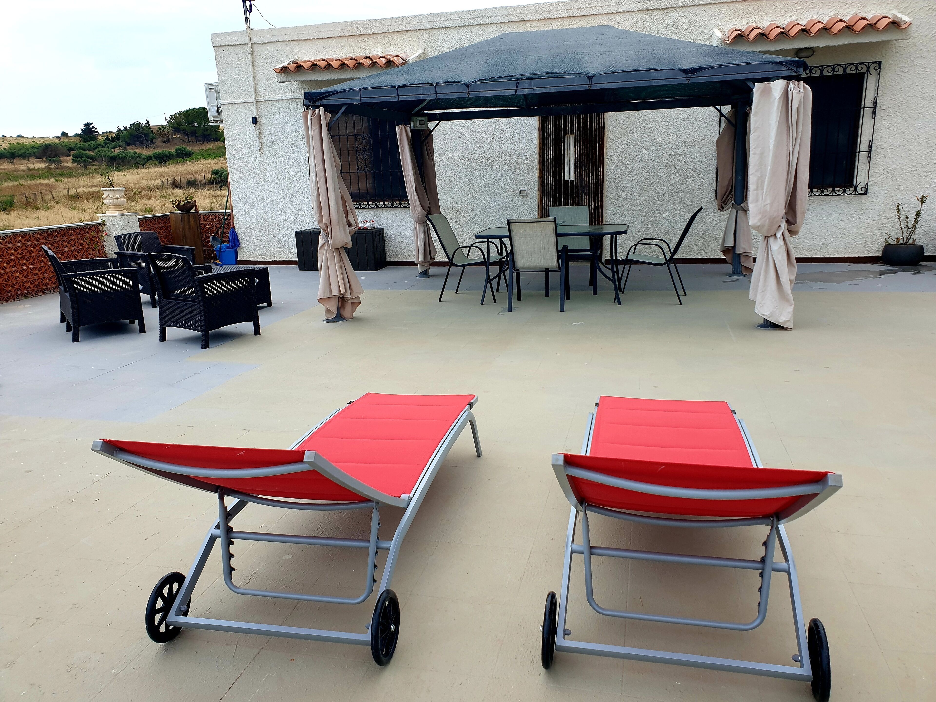 Una terrazza esterna con due poltrone rosse, una zona relax coperta con sedie e un tavolo, e un edificio bianco con tetto in tegole sullo sfondo.