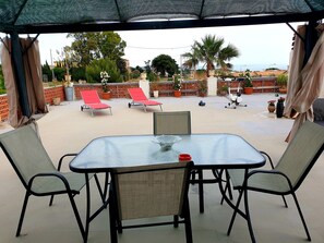 Un espace extérieur aménagé, abrité et doté de tables et de chaises, offrant une vue sur un jardin paysager agrémenté de palmiers et de plantes en pot.
