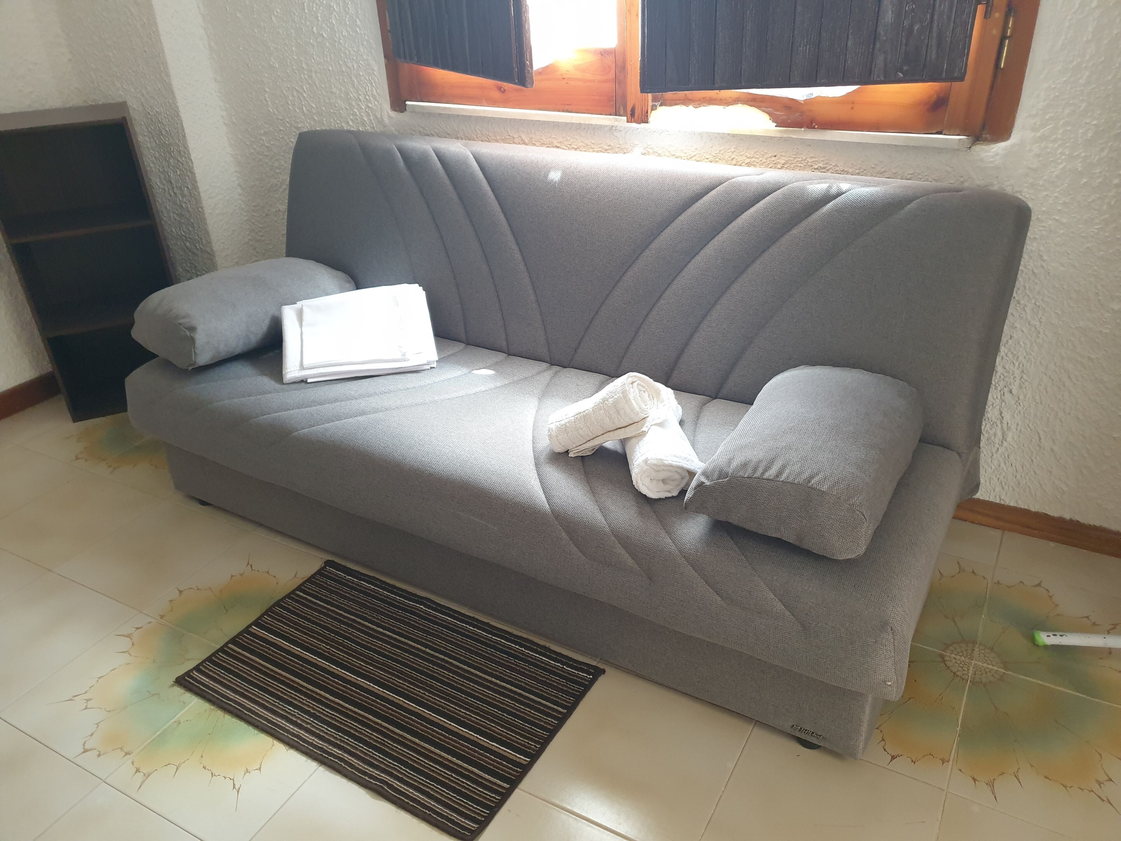 A grey sofa with a rolled-up white towel and a white pillow, a black mat in front, and a window with blinds.