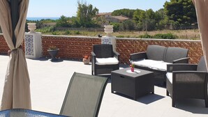 A covered outdoor seating area with wicker furniture, potted plants, and a view of the sea. - Mini apartment "CirPi" (max 4 people) Desusino near the sea (Butera)