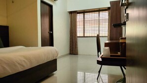 A hotel room with a bed, a desk, a chair, a radiator, and a window with curtains. - New Harshita inn By Glitz Hotel  (Navi Mumbai)