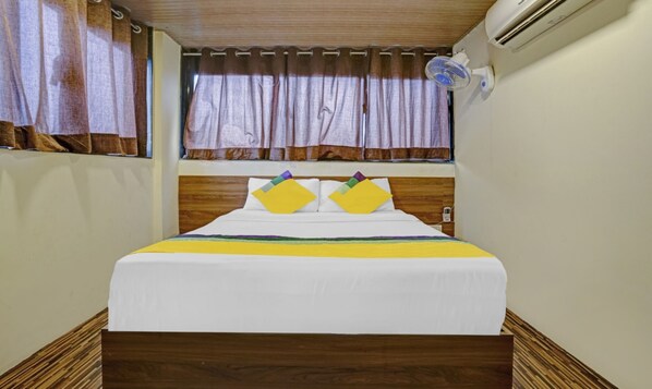 Une chambre d’hôtel avec une tête de lit en bois, un lit avec une literie blanche et des oreillers colorés, une fenêtre avec des rideaux et un climatiseur fixé au mur.