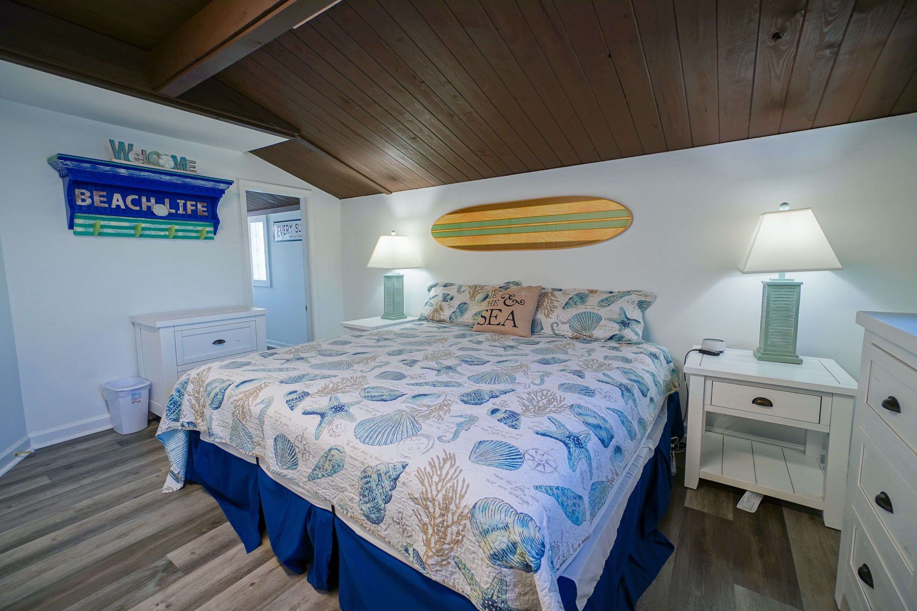 A bedroom with a bed, nightstands, a lamp, a trash can, and a 'Welcome to Beach Life' sign.