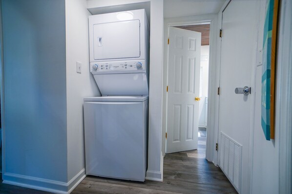 Une buanderie équipée d’un lave-linge et d’un sèche-linge superposés, avec un plancher en bois et des murs blancs.