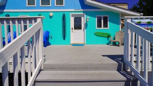 A turquoise house with a white door, a porch with a wooden railing, and a green surfboard leaning against the wall.