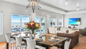 A spacious dining area with a large table set for six, surrounded by white chairs. A modern chandelier hangs above, and a large window offers a view of the ocean and boats.