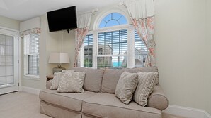 Ett vardagsrum med en beige soffa, en platt-TV som är monterad på väggen och ett fönster med dekorativa gardiner.