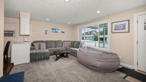 A living room with a grey sofa, a coffee table, a bean bag, and a window with a view of houses.