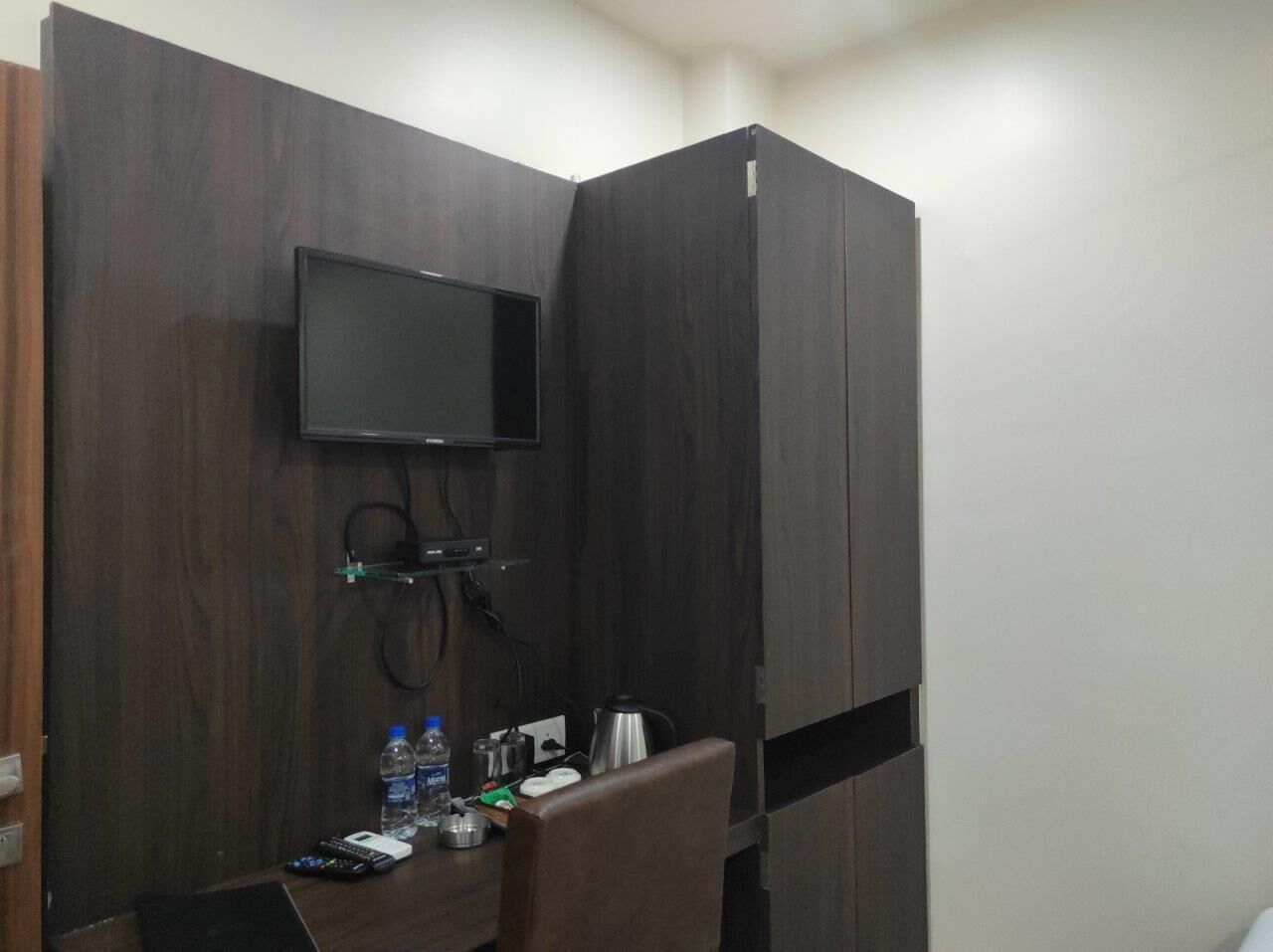 A small room with a dark wood wardrobe, a mounted TV, a desk with a chair, and a shelf with toiletries.