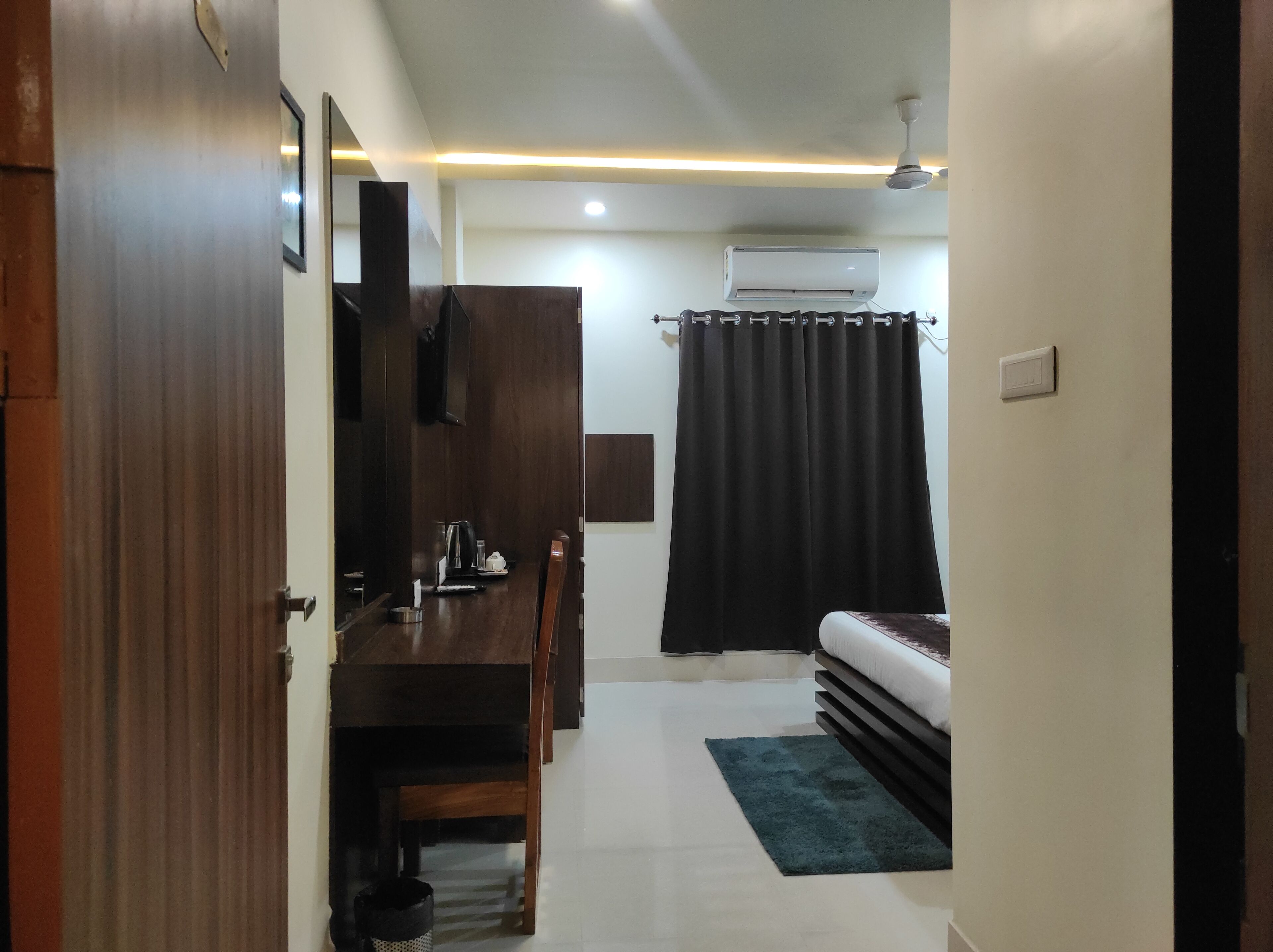 A compact room with a wooden desk, a bed, a dark curtain, and an air conditioning unit.