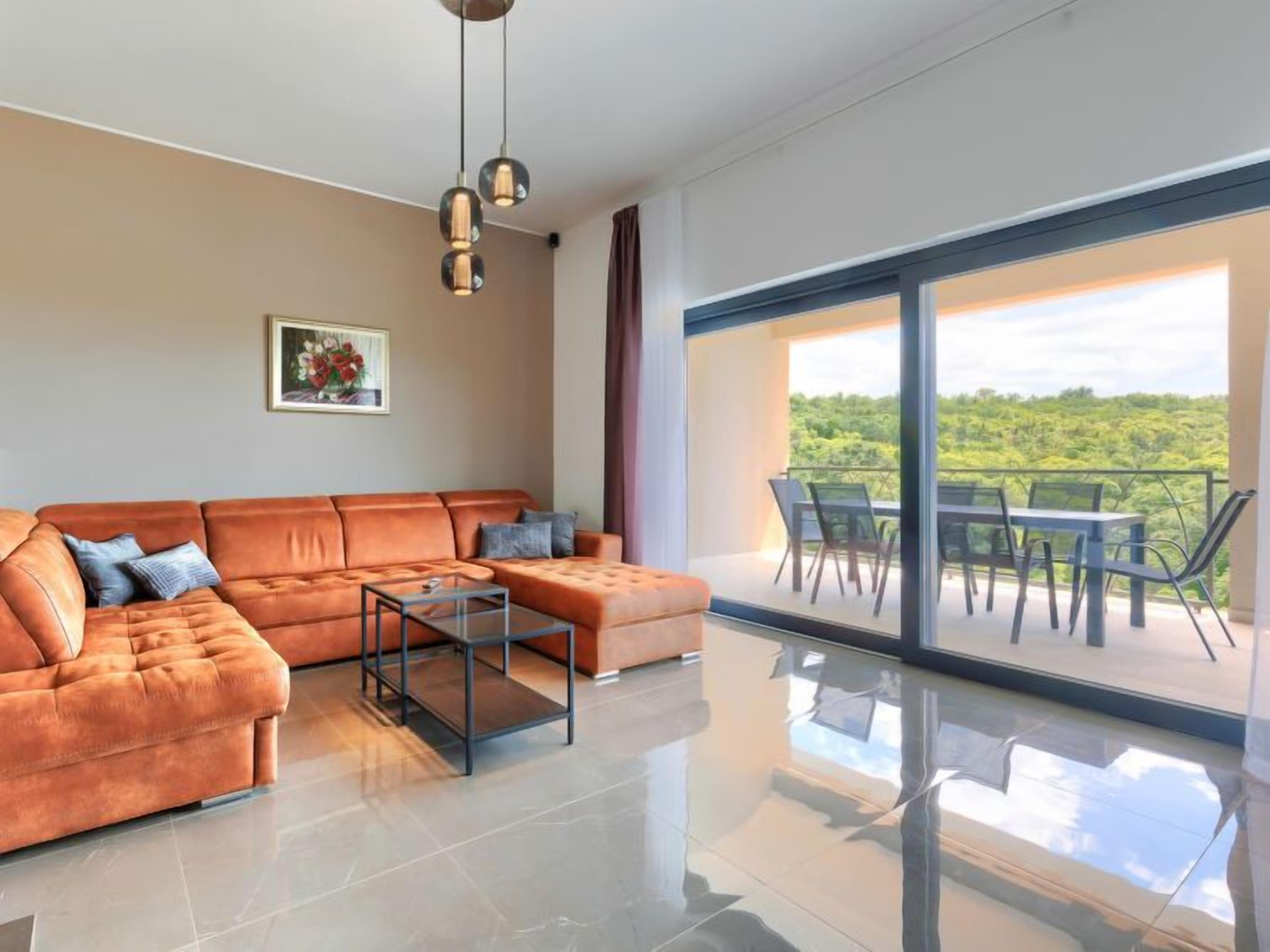 A modern living room with a leather sofa, a glass coffee table, and a large sliding glass door leading to an outdoor dining area with a view of greenery.