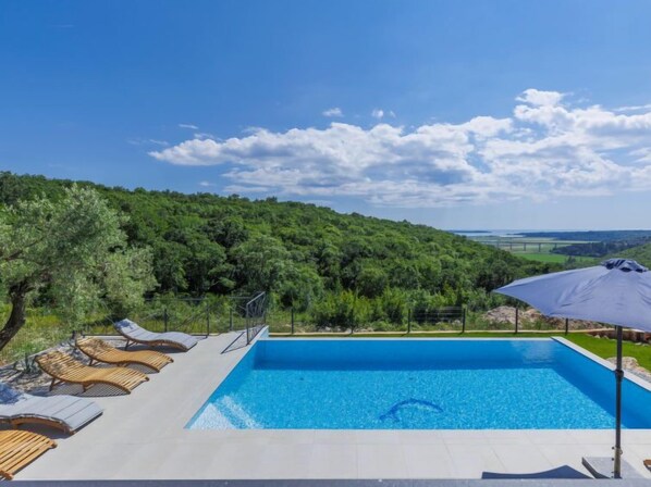 Ein rechteckiger Swimmingpool mit klar blauem Wasser, umgeben von einer gefliesten Liegefläche mit Holzliegen und einem großen Sonnenschirm.