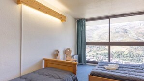Een slaapkamer met twee bedden, een houten hoofdeinde, een nachtkastje en een groot raam met uitzicht op een bergachtig landschap.