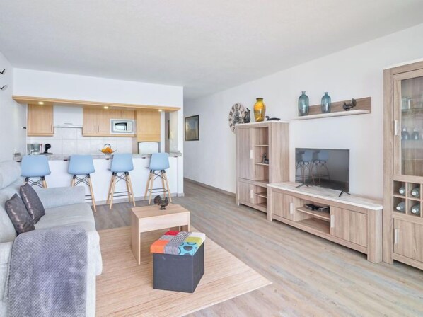 A modern living room with a sofa, a coffee table, and a TV. There is a kitchen area with a microwave and a sink.