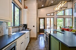A modern kitchen with a large island, wooden cabinets, and a view of the forest through large windows.