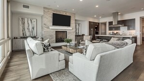 A modern living room with a fireplace, a flat-screen TV mounted above it, and a kitchen area with dark cabinets and stainless steel appliances. - Premium Five Bedroom Suite 5 Apartment Hotel by Redawning (Park City)
