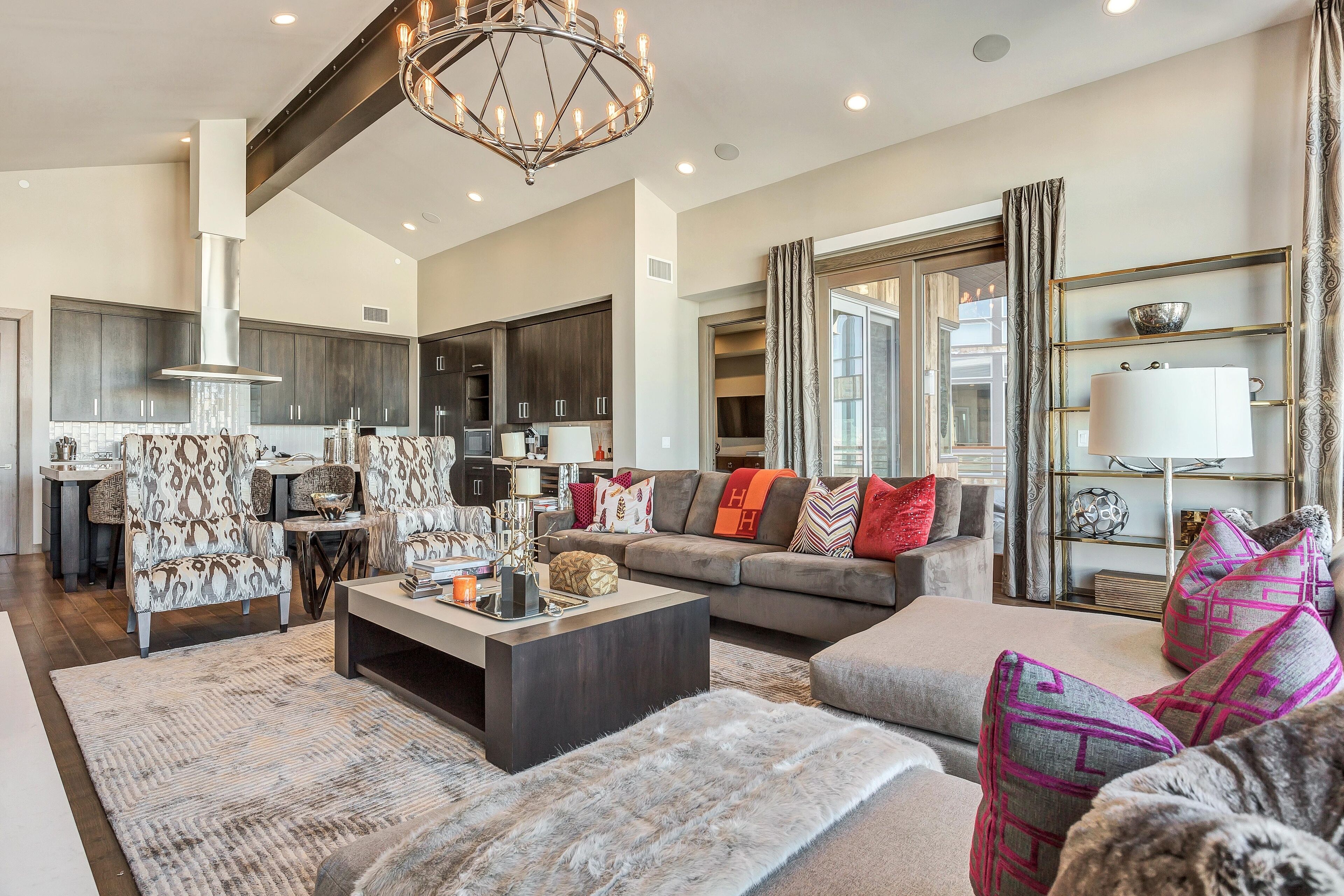 A spacious living room with a large sofa, a coffee table, and patterned armchairs. A kitchen with dark cabinets and a chimney is visible in the background.