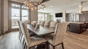A spacious dining area with a large wooden table and upholstered chairs, a modern kitchen with a built-in island, and large windows offering a view of the outdoors.