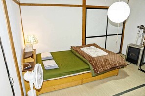 A single bed with a green bedspread and two pillows, a bedside table with a lamp, a white fan, and a heater on the right. - Pet friendly private lodge OMOTENASHI LODGE　YOUYOU (空知郡上富良野町)