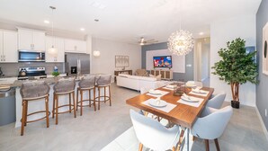 Una cocina moderna con armarios blancos, un comedor con mesa de madera y taburetes de bar, y una sala de estar con sofá y televisor.