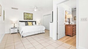 Une chambre à coucher comprenant un lit, des tables de chevet, un ventilateur de plafond, une porte donnant sur une autre pièce et une commode en bois.