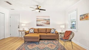 Un salon avec un canapé d’angle marron, un tapis à motifs, un ventilateur de plafond, un tableau accroché au mur et une fenêtre avec des stores.