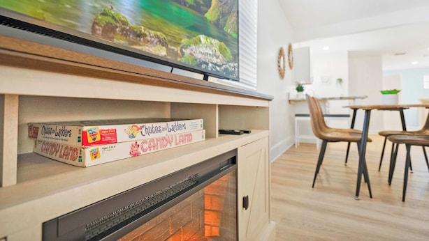 Un salon moderne avec un téléviseur à écran plat fixé au-dessus d’une cheminée, un coin repas avec des chaises en bois et une cuisine en arrière-plan.