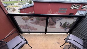 Ein Balkon mit zwei Metallstühlen und einem Tisch, der auf ein rotes Gebäude und ein grünes Feld blickt.