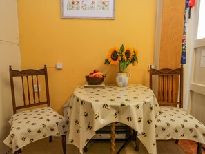 A dining area with a yellow wall, a framed floral artwork, a table with a bee-patterned tablecloth, a bowl of fruit, and a vase with sunflowers. - 2 Gateside Cottages (Coniston)