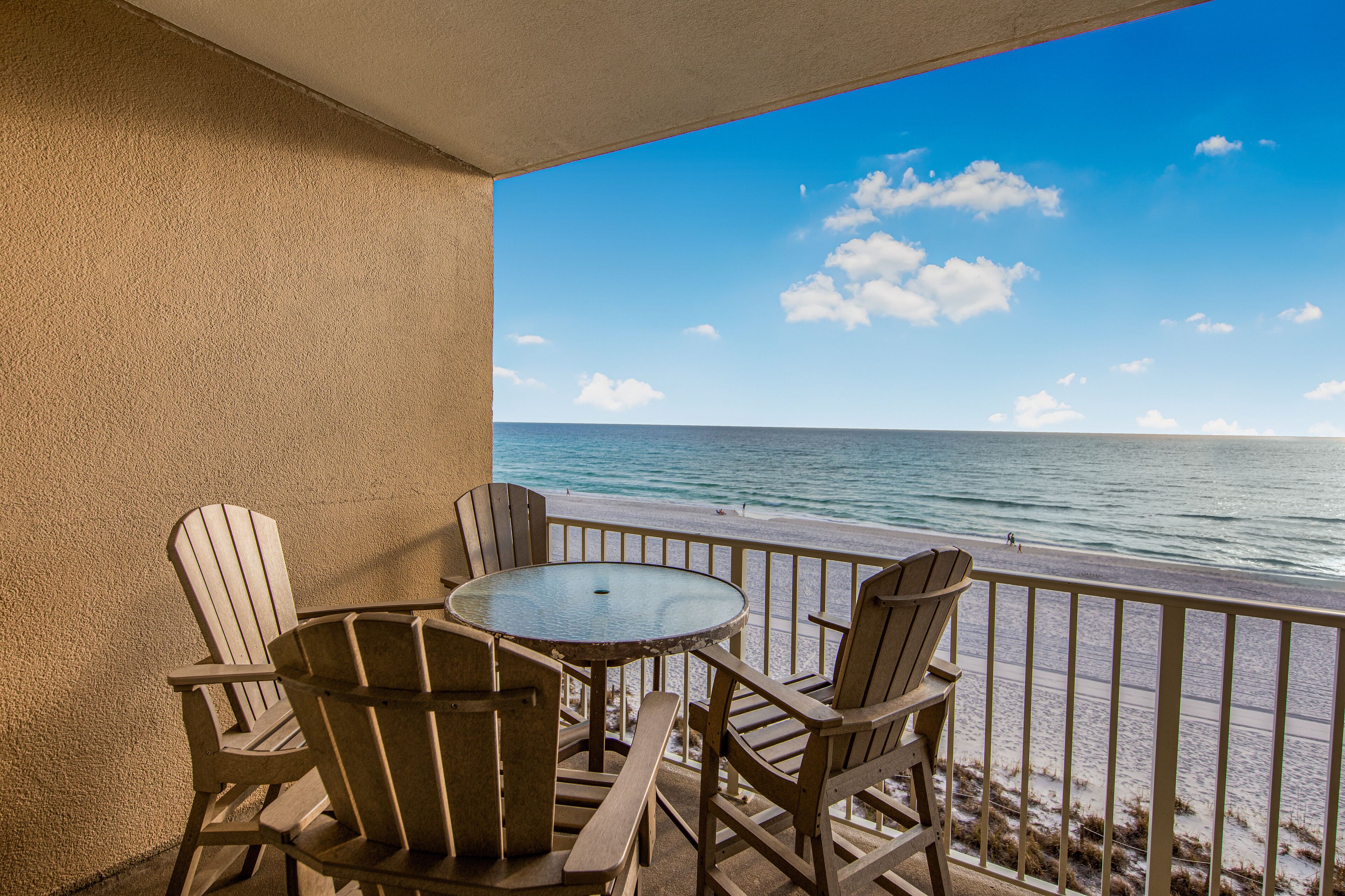 A balcony with a round table and four wooden chairs, overlooking a beach with the ocean and a clear sky.