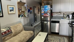 Un espace de vie compact comprenant un canapé, une petite table, un réfrigérateur, un lave-vaisselle et un coin cuisine avec des placards et un plan de travail.