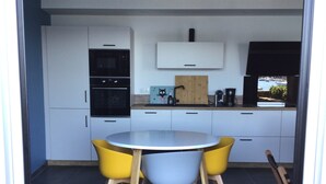 A modern kitchen with white cabinets, a black oven, a microwave, a wooden cutting board, and a coffee maker. There is a round table with four chairs in front of the kitchen. - Location Saisonnière Île Tudy « Sur Leau » (Île-Tudy)
