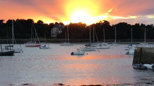 A marina with multiple boats docked, a large house in the background, and a sunset over the water. - Location Saisonnière Île Tudy « Sur Leau » (Île-Tudy)
