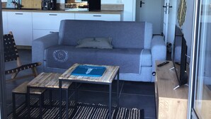 A compact living space with a sofa, a small table, a TV, and a kitchenette. - Location Saisonnière Île Tudy « Sur Leau » (Île-Tudy)