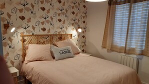 A bedroom with a bed, a nightstand, a wall-mounted lamp, and a window with curtains. - Location Saisonnière Île Tudy « Sur Leau » (Île-Tudy)