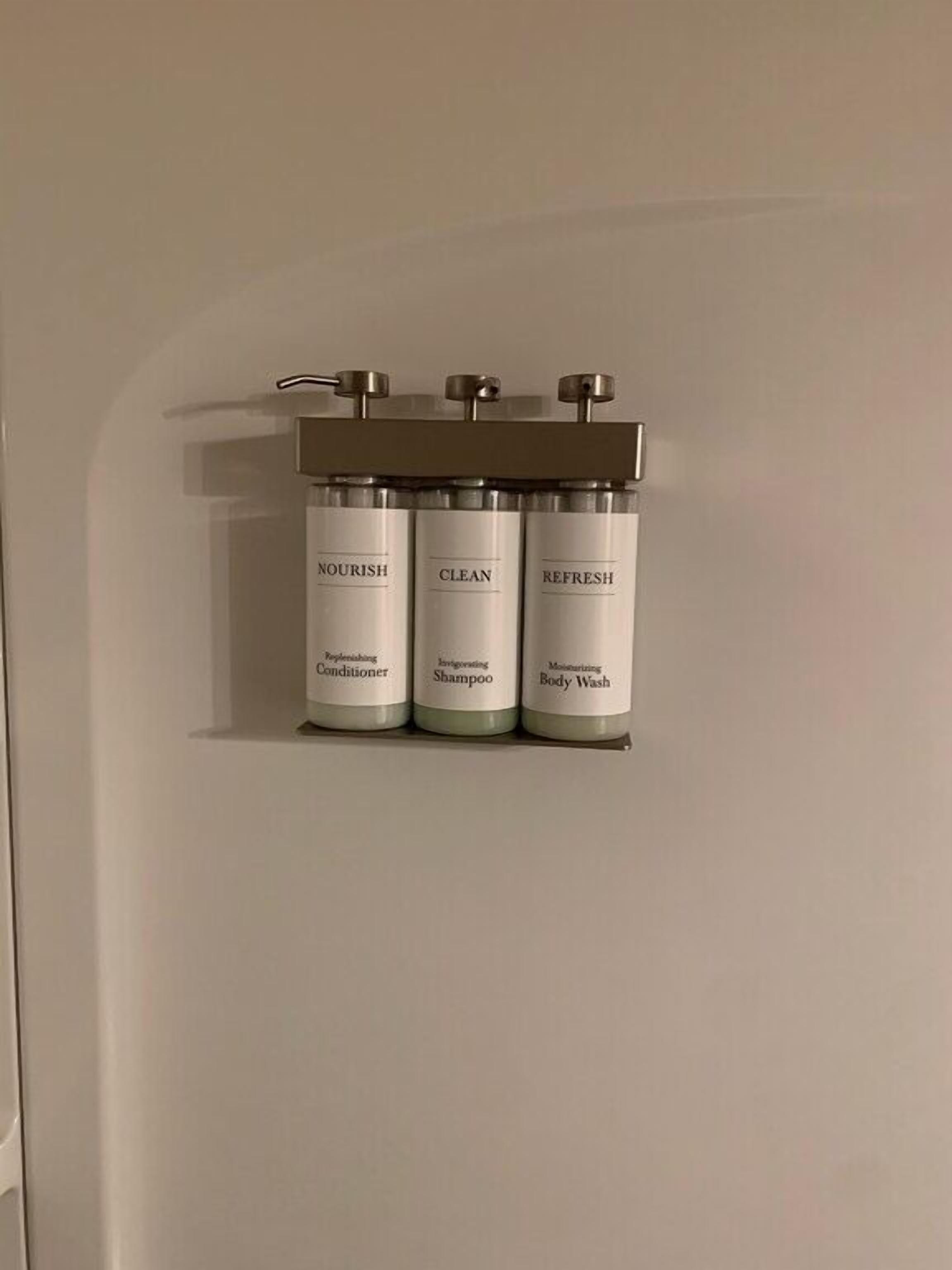 Ein Badezimmer mit einem wandmontierten Spender, auf dem drei Flaschen mit Toilettenartikeln mit den Aufschriften 'Nourish', 'Clean' und 'Refresh' stehen.