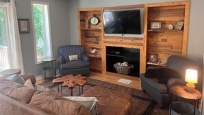 Ein Wohnzimmer mit Deckenventilator, Fernseher an einer Holzwand, Kamin und großem Fenster.