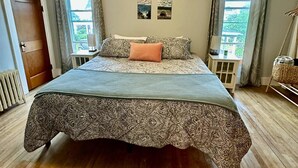 A bedroom with a large bed, wooden flooring, a ceiling light, and four wall-mounted pictures.