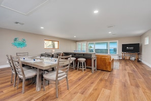 A spacious living area with a dining table, chairs, a kitchen counter, a TV, and a view of the ocean.