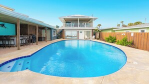 A swimming pool with a wooden deck, a two-story building with a balcony, and a fenced yard.