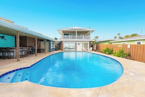 En swimmingpool med træterrasse, en toetagers bygning med balkon, og en indhegnet have.