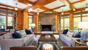 A spacious living room with a stone fireplace, wooden beams, and large windows offering a view of the outdoors. - Gorgeous 4 bedroom ski-in/out condominium, immaculate interior!! (Bachelor Gulch)
