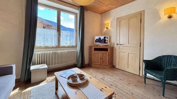 A cozy room with a wooden floor, a wooden table with a tray, a green chair, a television on a wooden cabinet, and a window with curtains. - Ground floor, tv, ski locker, 42m², Praz sur Arly (Praz-sur-Arly)