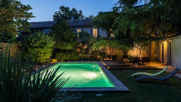 Una piscina con un bordeado, un patio con mobiliario exterior y una casa al fondo.