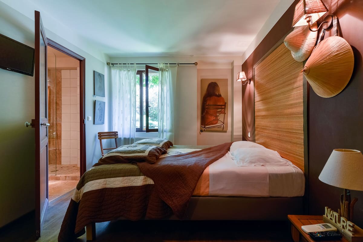 A hotel room with a bed, a chair, a desk with a lamp, and a painting on the wall.